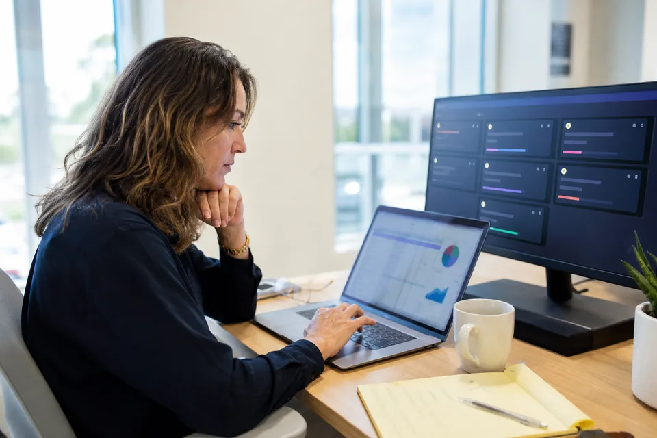 Engineering manager comparing coding agent seat costs on a laptop next to a monitor with agent task panels